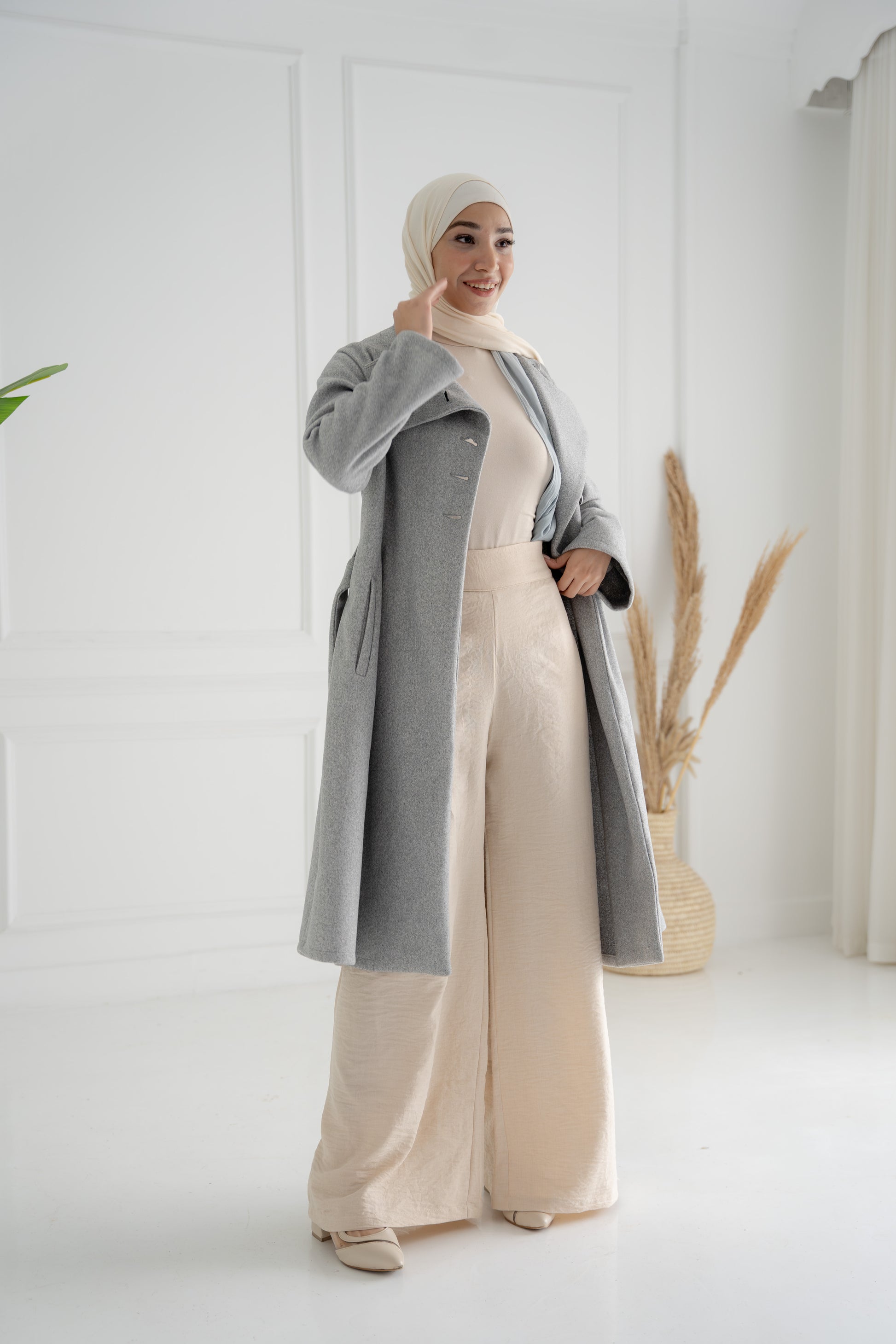 A model showcasing the Misty Gray Coat, a long, elegant coat made of fine broadcloth fabric. The coat features a waist belt, side pockets, and a unique button detail across the chest. Styled with wide pants, this versatile piece is perfect for both casual and formal settings.