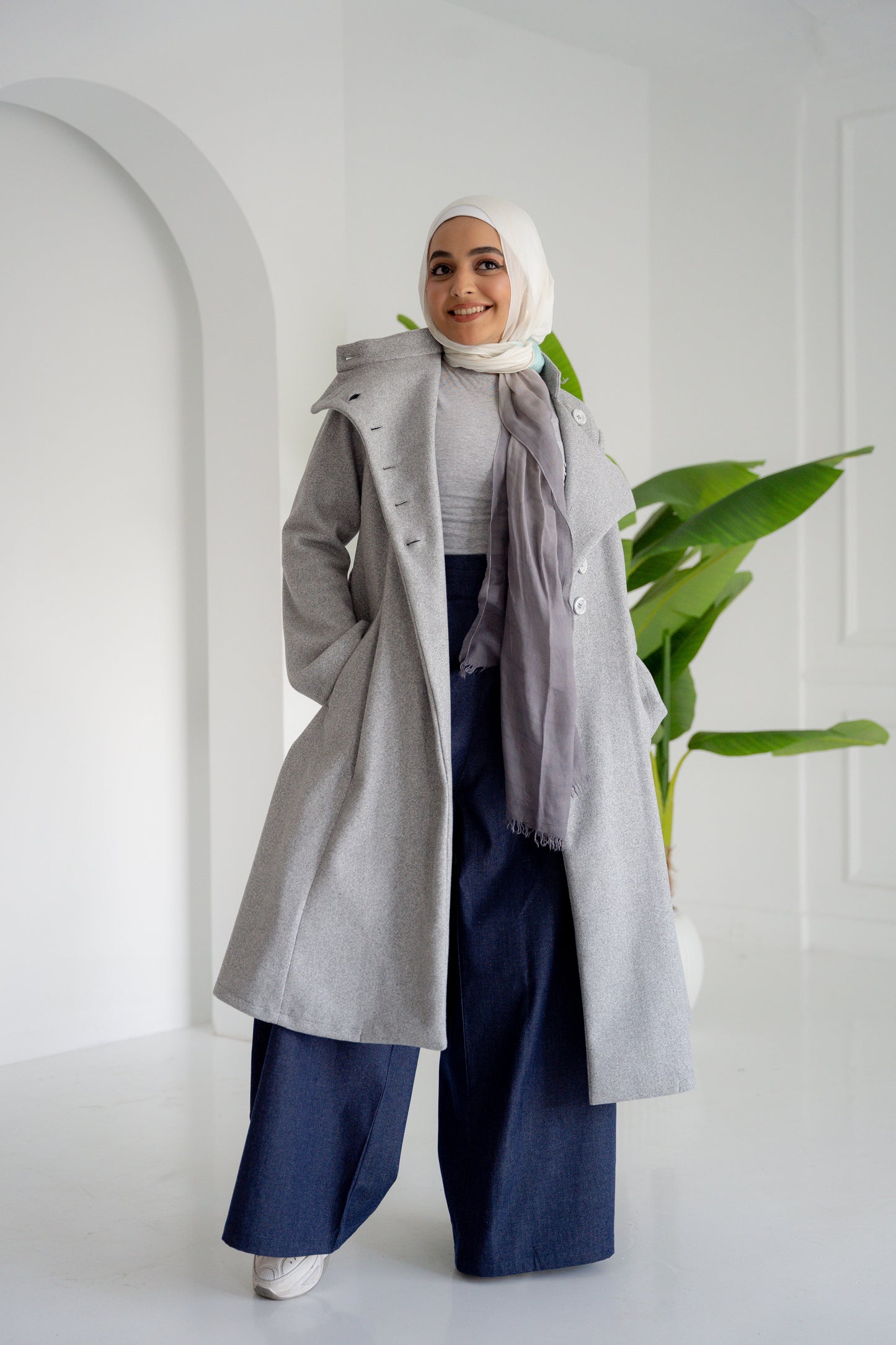 A model showcasing the Misty Gray Coat, a long, elegant coat made of fine broadcloth fabric. The coat features a waist belt, side pockets, and a unique button detail across the chest. Styled with wide pants, this versatile piece is perfect for both casual and formal settings.