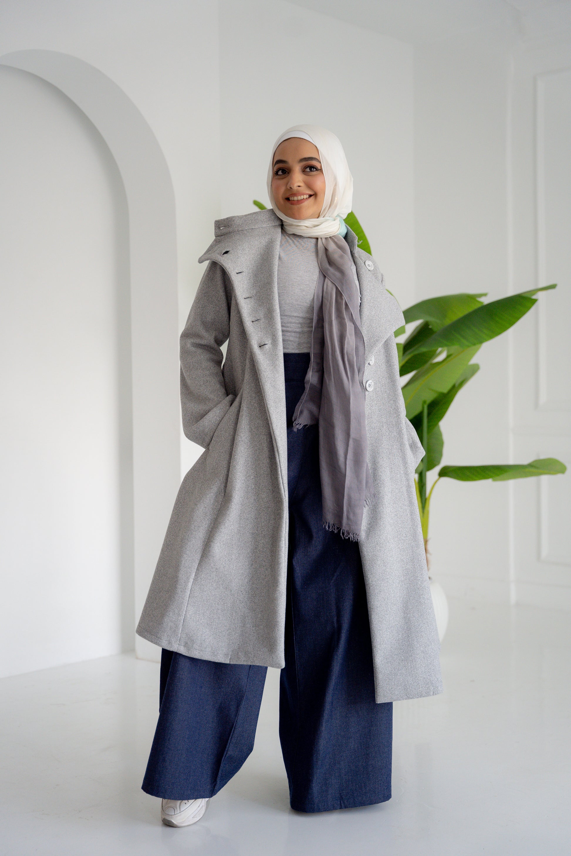 A model showcasing the Misty Gray Coat, a long, elegant coat made of fine broadcloth fabric. The coat features a waist belt, side pockets, and a unique button detail across the chest. Styled with wide pants, this versatile piece is perfect for both casual and formal settings.