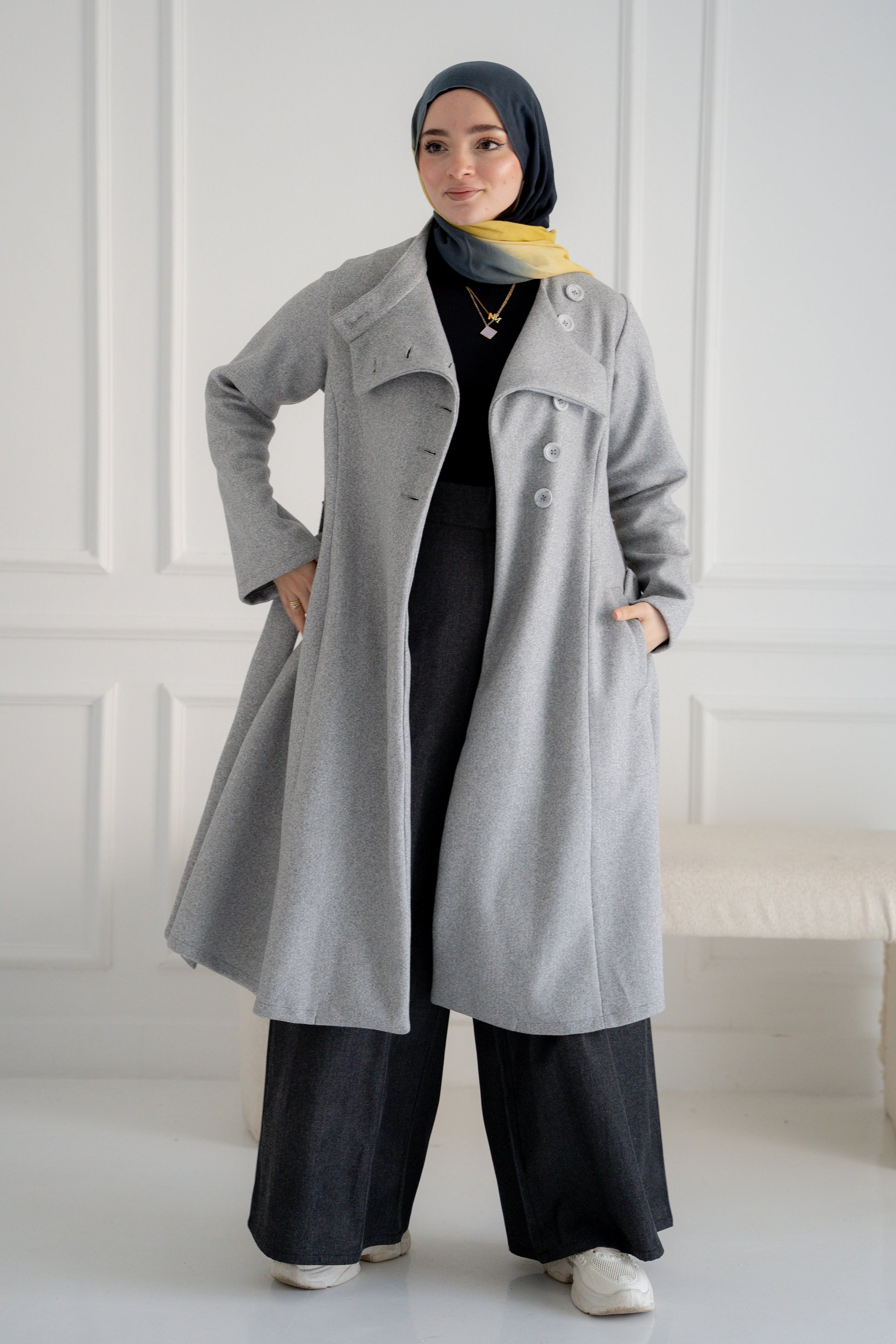 A model showcasing the Misty Gray Coat, a long, elegant coat made of fine broadcloth fabric. The coat features a waist belt, side pockets, and a unique button detail across the chest. Styled with wide pants, this versatile piece is perfect for both casual and formal settings.