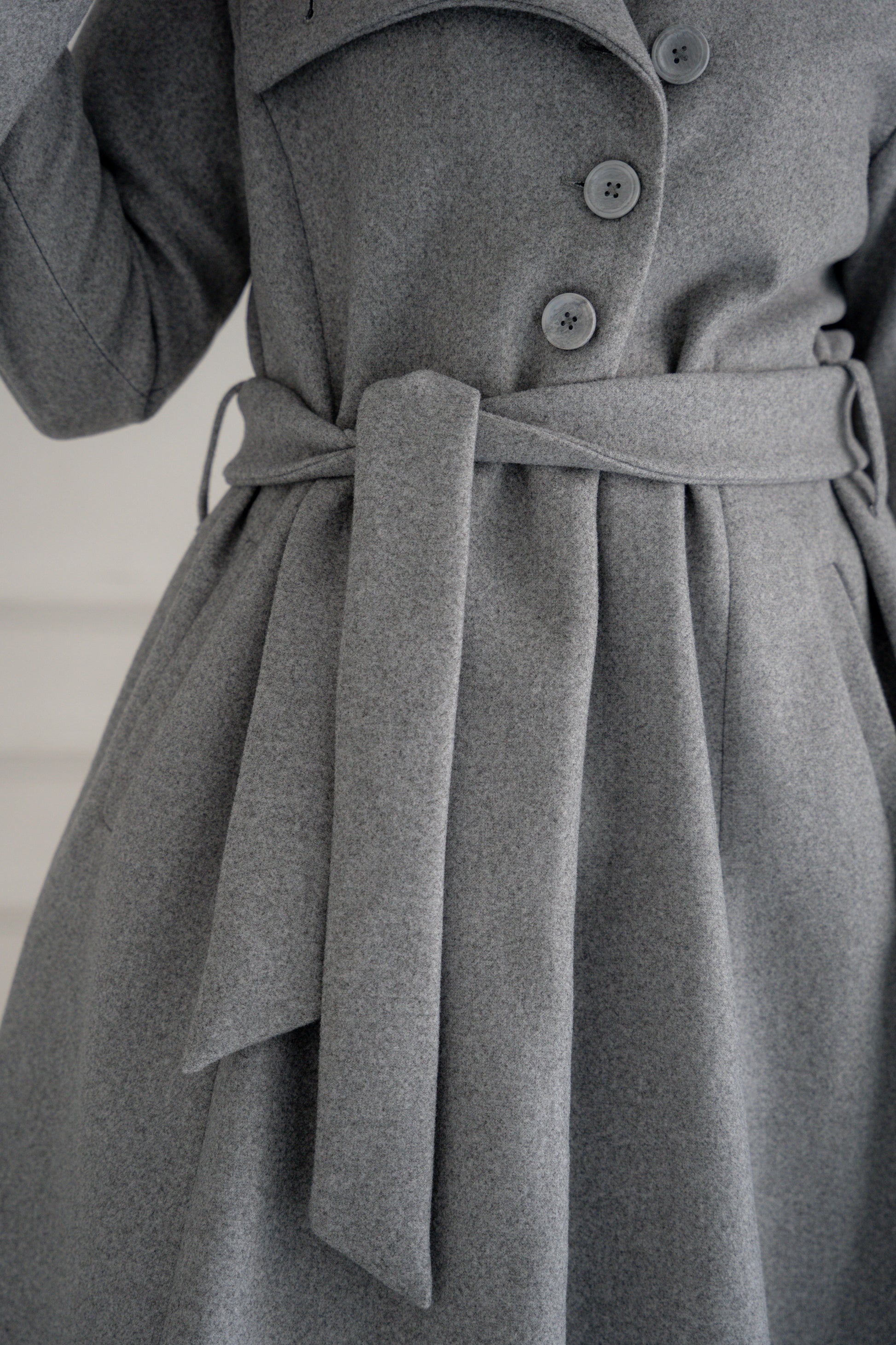 A model showcasing the Misty Gray Coat, a long, elegant coat made of fine broadcloth fabric. The coat features a waist belt, side pockets, and a unique button detail across the chest. Styled with wide pants, this versatile piece is perfect for both casual and formal settings.